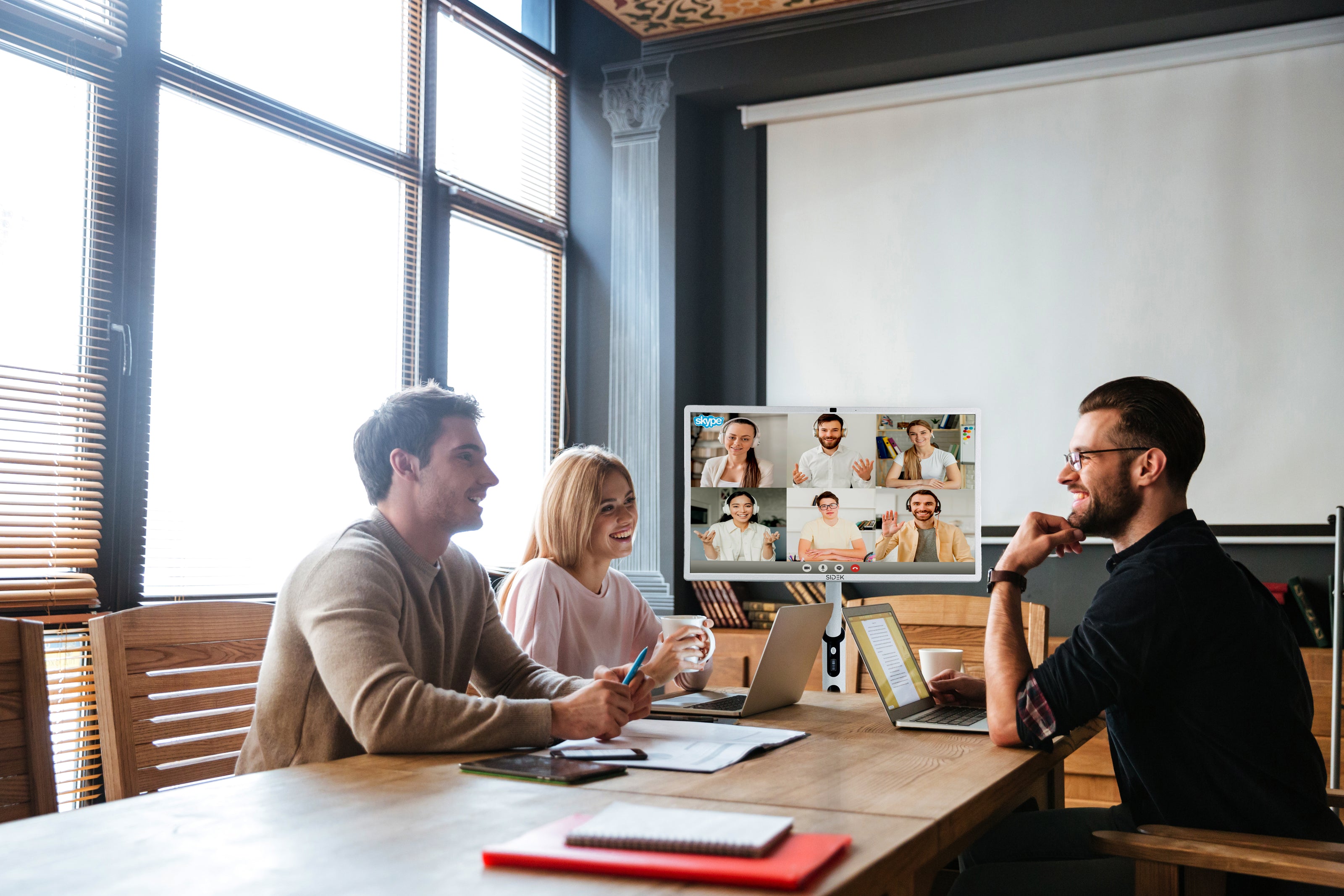 a group of professionals in a modern office space engaged in a video conference call displayed on a sidek itv smart display. the screen shows multiple remote participants, enhancing collaboration during the meeting. the setting features natural lighting, wooden furniture, and an interactive workspace.