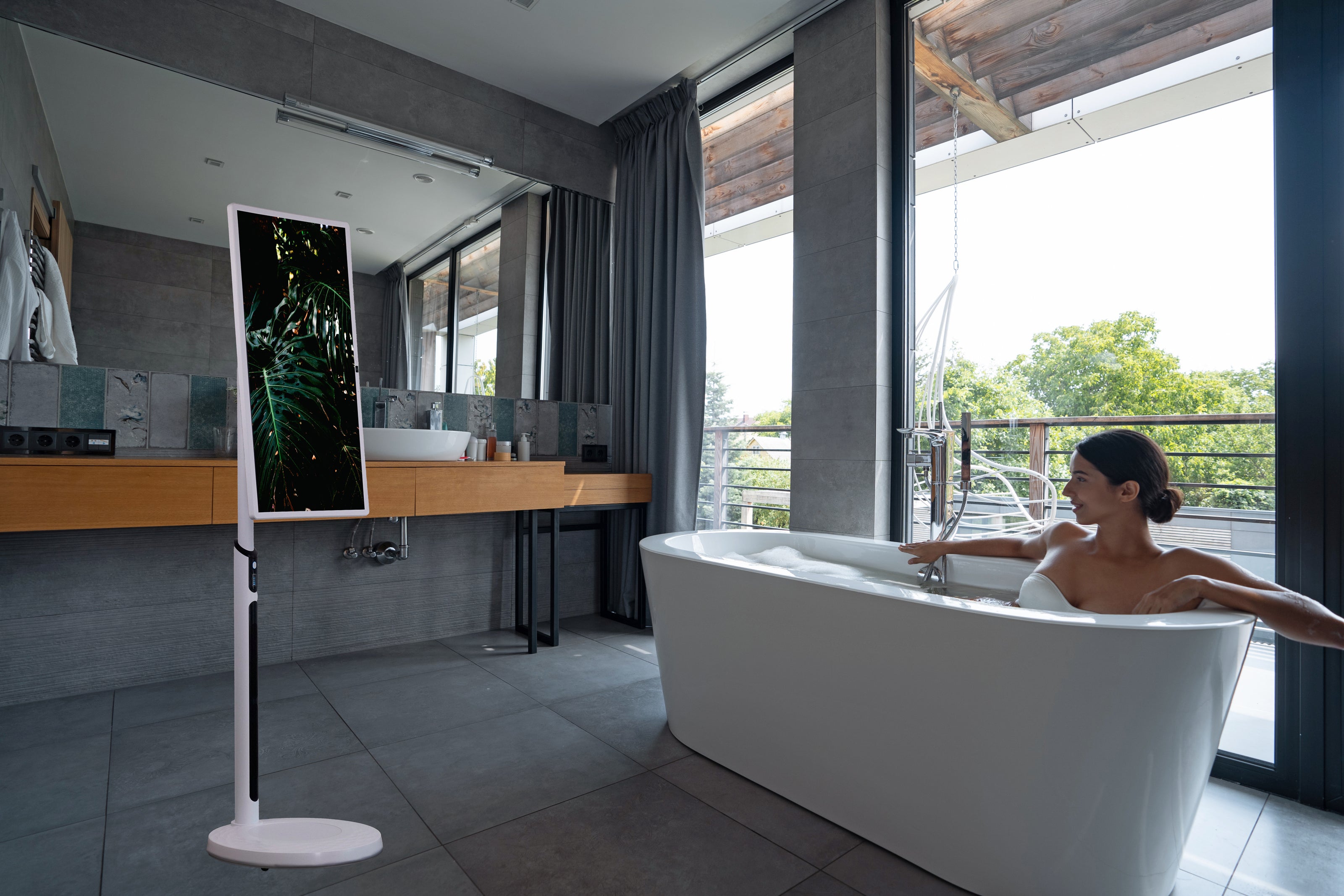 a woman relaxing in a modern bathtub while watching content on a sidek itv smart display. the sleek, freestanding screen is positioned in a stylish bathroom with large windows, providing an immersive viewing experience in a luxurious setting.